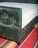 A luxurious green leather upholstered bed, American-made with clean lines and hand burnished details. Features unique nickel nail trim with cactus designs on the headboard and footboard, presenting a tall, art-like stature in the bedroom.
