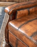 A brown leather recliner chair with a wooden base and armrests is pictured against a plain beige background. The chair has a tight back with a boot stitch emblem and is adorned with premium upholstery grade brindle cowhide on the sides and outside back. T