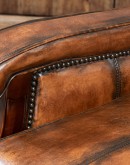 A brown leather recliner chair with a wooden base and armrests is pictured against a plain beige background. The chair has a tight back with a boot stitch emblem and is adorned with premium upholstery grade brindle cowhide on the sides and outside back. T