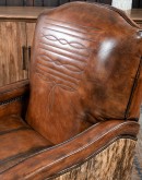 A brown leather recliner chair with a wooden base and armrests is pictured against a plain beige background. The chair has a tight back with a boot stitch emblem and is adorned with premium upholstery grade brindle cowhide on the sides and outside back. T