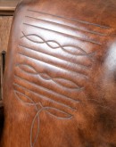 A brown leather recliner chair with a wooden base and armrests is pictured against a plain beige background. The chair has a tight back with a boot stitch emblem and is adorned with premium upholstery grade brindle cowhide on the sides and outside back. T