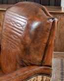 A brown leather recliner chair with a wooden base and armrests is pictured against a plain beige background. The chair has a tight back with a boot stitch emblem and is adorned with premium upholstery grade brindle cowhide on the sides and outside back. T