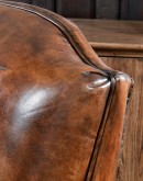 A brown leather recliner chair with a wooden base and armrests is pictured against a plain beige background. The chair has a tight back with a boot stitch emblem and is adorned with premium upholstery grade brindle cowhide on the sides and outside back. T