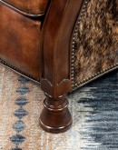 A brown leather recliner chair with a wooden base and armrests is pictured against a plain beige background. The chair has a tight back with a boot stitch emblem and is adorned with premium upholstery grade brindle cowhide on the sides and outside back. T