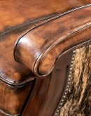 A brown leather recliner chair with a wooden base and armrests is pictured against a plain beige background. The chair has a tight back with a boot stitch emblem and is adorned with premium upholstery grade brindle cowhide on the sides and outside back. T