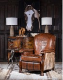 A brown leather recliner chair with a wooden base and armrests is pictured against a plain beige background. The chair has a tight back with a boot stitch emblem and is adorned with premium upholstery grade brindle cowhide on the sides and outside back. T