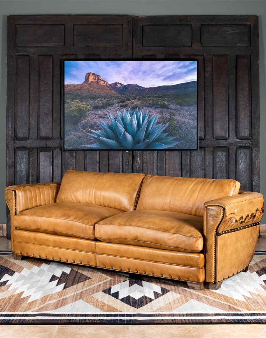 A luxurious tan leather sofa with hand-painted horses and canyon scenes by artist Wayne Parmenter, featuring classic cowboy boot stitching and channel design on the arms, part of Adobe Interiors' Desert Ranch and Mountain Ranch Collection.