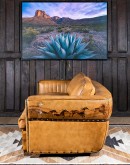 A luxurious tan leather sofa with hand-painted horses and canyon scenes by artist Wayne Parmenter, featuring classic cowboy boot stitching and channel design on the arms, part of Adobe Interiors' Desert Ranch and Mountain Ranch Collection.