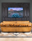 A luxurious tan leather sofa with hand-painted horses and canyon scenes by artist Wayne Parmenter, featuring classic cowboy boot stitching and channel design on the arms, part of Adobe Interiors' Desert Ranch and Mountain Ranch Collection.