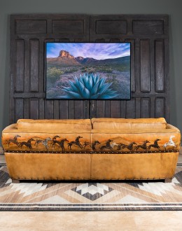 A luxurious tan leather sofa with hand-painted horses and canyon scenes by artist Wayne Parmenter, featuring classic cowboy boot stitching and channel design on the arms, part of Adobe Interiors' Desert Ranch and Mountain Ranch Collection.