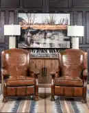 A brown leather recliner chair with a wooden base and armrests is pictured against a plain beige background. The chair has a tight back with a boot stitch emblem and is adorned with premium upholstery grade brindle cowhide on the sides and outside back. T
