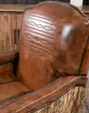 A brown leather recliner chair with a wooden base and armrests is pictured against a plain beige background. The chair has a tight back with a boot stitch emblem and is adorned with premium upholstery grade brindle cowhide on the sides and outside back. T