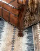 A brown leather recliner chair with a wooden base and armrests is pictured against a plain beige background. The chair has a tight back with a boot stitch emblem and is adorned with premium upholstery grade brindle cowhide on the sides and outside back. T