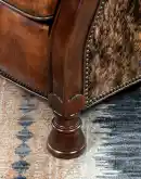 A brown leather recliner chair with a wooden base and armrests is pictured against a plain beige background. The chair has a tight back with a boot stitch emblem and is adorned with premium upholstery grade brindle cowhide on the sides and outside back. T